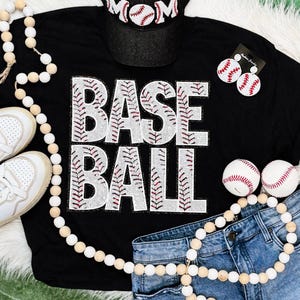 May include: Black baseball-themed t-shirt with the word "BASEBALL" in white, baseball-stitch-style lettering. Accessories include a baseball cap with "MOM" text, baseball earrings, two baseballs, and a pair of jeans.