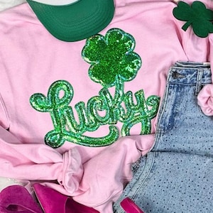 May include: A pink sweatshirt with a large green glitter clover and the word "Lucky" in green glitter. A green baseball cap, denim shorts, and pink shoes are also visible.
