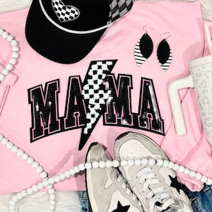 May include: Pink t-shirt with a black and white checkered lightning bolt graphic and the word "MAMA" in black with pink outlining. The shirt is paired with a black and white checkered trucker hat, white beaded necklace, and white sneakers with black stars.