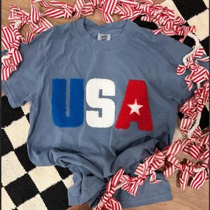May include: A dusty blue short-sleeve t-shirt featuring the letters USA in large, fuzzy, textured chenille patches. The U is blue, the S is white, and the A is red with a small white star centered on the letter. The shirt has a crew neckline.