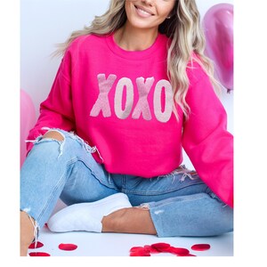 May include: A bright pink sweatshirt with the word "XOXO" in a lighter pink, fuzzy font. The sweatshirt is paired with light blue ripped jeans and white sneakers. Red rose petals are scattered on the floor.
