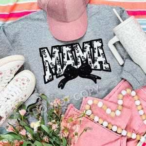May include: A gray sweatshirt with the word "MAMA" in black and white floral print. The sweatshirt is laid out on a pink and white striped blanket with a pink hat, a white tumbler, and pink flowers.