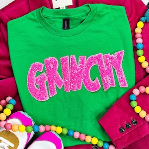 Puede incluir: Una camiseta verde con la palabra "GRINCHY" en letras rosas y brillantes. La camiseta se muestra con una blazer rosa, collares de cuentas coloridas y zapatillas rosas y blancas.
