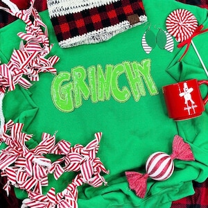 Puede incluir: Una sudadera verde con la palabra "GRINCHY" en letras con lentejuelas. La imagen incluye una taza roja con un gráfico blanco, aretes a rayas, un bastón de caramelo y un gorro con un patrón de cuadros rojos y negros. Arcos a rayas rojas y blancas están dispersos.
