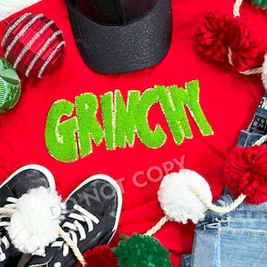 Puede incluir: Una camiseta roja con la palabra "GRINCH" en letras verdes y brillantes. La imagen también incluye una gorra de béisbol negra, zapatillas negras y adornos navideños. También es visible una guirnalda de pompones rojos y blancos.