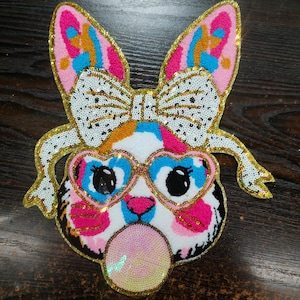 May include: A colourful sequined iron-on patch featuring a cartoon animal with pink and blue heart-shaped sunglasses, a white bow, and blowing a bubble. The animal has a pink, blue, and white patterned face with black eyes.
