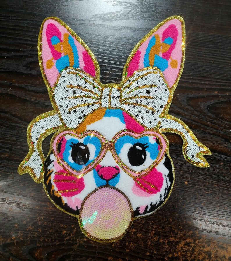 May include: A colourful sequined iron-on patch featuring a cartoon animal with pink and blue heart-shaped sunglasses, a white bow, and blowing a bubble. The animal has a pink, blue, and white patterned face with black eyes.