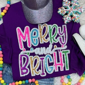 May include: A purple sweatshirt with the words "Merry and Bright" in colorful sequined letters. A silver glitter baseball cap and a snowflake ornament are visible. The sweatshirt is paired with blue jeans and white shoes.