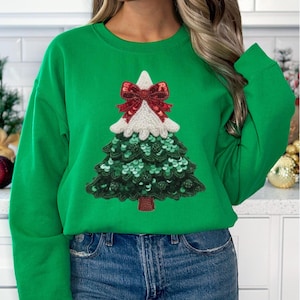 May include: A green sweatshirt featuring a sequined Christmas tree design. The tree has a white top, a red bow, and green sequin details. The sweatshirt is a solid, bright green color.