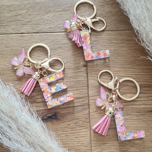Könnte beinhalten: Drei Schlüsselanhänger mit goldenen Ringen und rosa Quasten. Jeder Schlüsselanhänger hat einen klaren Buchstaben-Anhänger, der mit buntem Glitzer gefüllt ist, und einen rosa Schmetterlings-Anhänger. Die Buchstaben sind "L", "E" und "L".