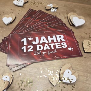Könnte beinhalten: Ein Stapel roter Karten mit weißem Text "1 Jahr 12 Dates Zeit zu zweit" ist von herzförmigen Holzdekorationen mit weißen Akzenten und goldenem Konfetti umgeben. Die Karten sind auf einer Holzoberfläche angeordnet.