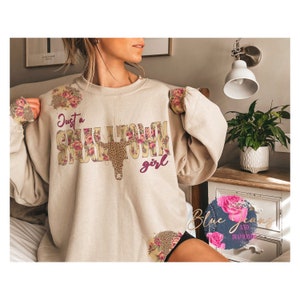 May include: A beige sweatshirt with the text "Just a Small Town Girl" in brown and pink lettering. The text is surrounded by a floral design and a brown and black cow skull.
