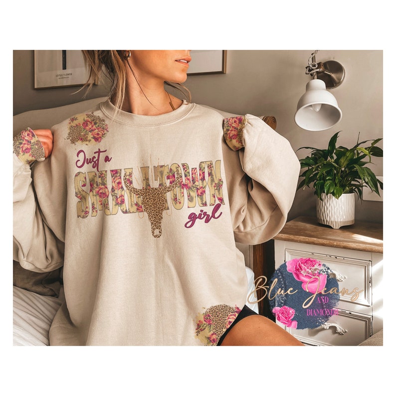 May include: A beige sweatshirt with the text "Just a Small Town Girl" in brown and pink lettering. The text is surrounded by a floral design and a brown and black cow skull.