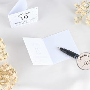 May include: A white card with black text that reads "Grund 19 Warum ich dich liebe". The card is folded in half and a black marker pen is lying on top of the card.