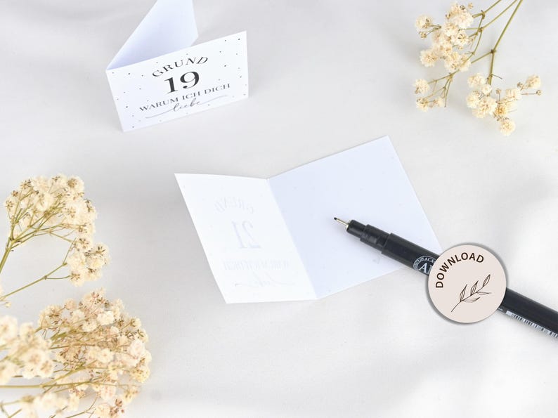 May include: A white card with black text that reads "Grund 19 Warum ich dich liebe". The card is folded in half and a black marker pen is lying on top of the card.