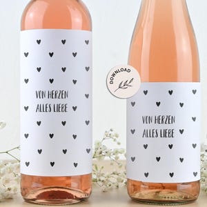 Pode incluir: Duas garrafas de vinho rosé com rótulos brancos com padrões de corações pretos e o texto "VON HERZEN ALLES LIEBE". Um autocolante "DOWNLOAD" está numa garrafa. Flores brancas no fundo.