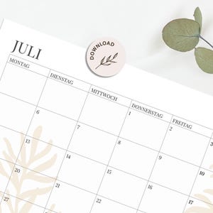 May include: A white monthly calendar page for the month of July with black text in German, featuring a grid layout with numbered days. A circular sticker with the text DOWNLOAD and a leaf illustration is placed on the top centre of the page.
