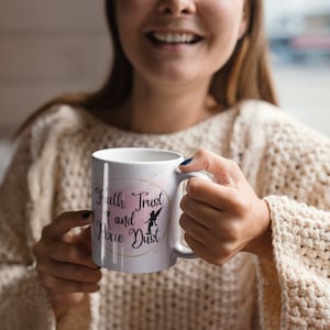 Puede incluir: Taza de cerámica blanca con el texto "Faith, Trust and Pixie Dust" en escritura negra, con un diseño rosa y dorado. La taza tiene un asa y la sostiene una persona que lleva un jersey color crema.