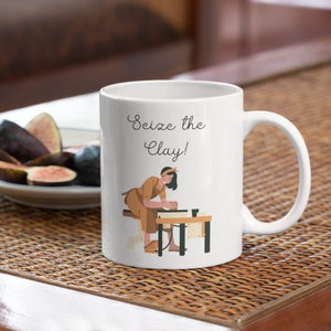 May include: White ceramic mug featuring the text "Seize the Clay!" and an illustration of a person at a pottery wheel. The person is dressed in brown clothing and a headband. The mug has a curved handle.