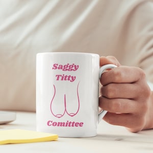 May include: White ceramic mug with the text "Saggy Titty Committee" in pink, along with a pink illustration of breasts. The mug is held by a person's hand.