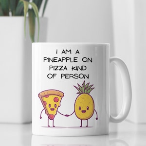 May include: White ceramic mug with the text "I AM A PINEAPPLE ON PIZZA KIND OF PERSON." The mug features cartoon illustrations of a pizza slice and a pineapple holding hands. The mug has a white handle.