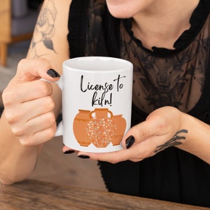 Puede incluir: Taza de cerámica blanca con el texto "License to kiln!" en escritura negra. La taza presenta un diseño de tres vasijas de cerámica de color terracota. La vasija central tiene un patrón de mosaico en tonos azules y naranjas.
