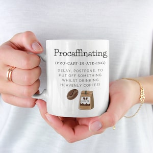Puede incluir: Taza de café de cerámica blanca con la palabra "Procaffinating" impresa en negro. La taza incluye una definición e ilustraciones de un grano de café y una bolsa de café. La taza está sostenida por una persona.