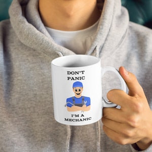 May include: White ceramic mug featuring the text "DON'T PANIC I'M A MECHANIC" and a cartoon mechanic graphic. The mechanic is in a blue uniform and hat, holding tools. The mug has a white handle.