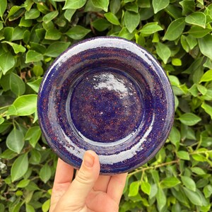 May include: A handmade, ceramic dish with a deep blue glaze and speckled red-brown accents. The dish has a rounded shape with a slightly raised rim. The pottery is held against a backdrop of green foliage.