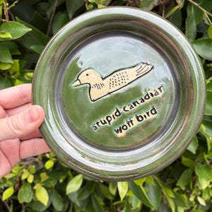 May include: A round, green ceramic dish with a glossy finish. The center features a light-colored, hand-drawn illustration of a bird and the text "stupid canadian wolf bird" etched into the surface in dark, lowercase lettering.