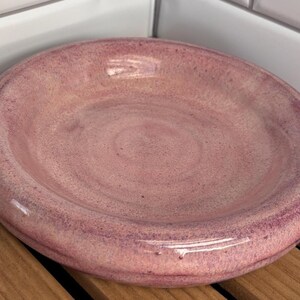 May include: A round, shallow ceramic bowl with a glossy, mottled pink and mauve glaze. The surface features subtle variations in color intensity, creating a textured appearance across the interior and the wide, rounded rim of the dish.