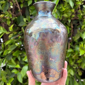 May include: A ceramic vase with a metallic, iridescent glaze featuring a crystalline pattern. The surface displays shifting hues of bronze, copper, blue, and purple, with a smooth, rounded body and a narrow, flared neck opening at the top.