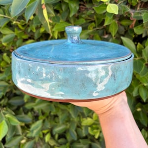 May include: A round, turquoise ceramic serving dish with a lid. The dish has a brown rim and a small handle on the lid. The turquoise glaze has a mottled appearance. The dish is held in front of a background of green leaves.