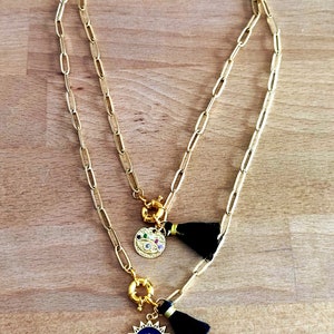 May include: A gold chain necklace with two charms. One charm is a gold circle with a blue eye and a black tassel. The other charm is a gold circle with a colorful eye and a black tassel.