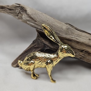 May include: A shiny gold-toned metal figurine of a hare in a standing position. The hare features textured detailing across its body to represent fur, with long, upright ears and a small tail. The surface of the metal is highly reflective.