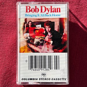 May include: A clear cassette tape of Bob Dylan's album "Bringing It All Back Home." The album cover features a photograph of Bob Dylan and a woman in a red dress. The cassette case has a barcode and the Columbia Stereo Cassette logo.