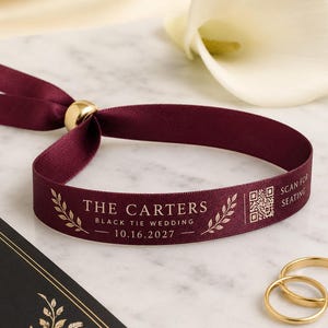 Pode incluir: Uma pulseira de tecido bordô profundo com um fecho de conta deslizante de metal dourado. A fita está impressa em dourado com o texto THE CARTERS, BLACK TIE WEDDING, 16.10.2027, SCAN FOR SEATING, um código QR e ilustrações de ramos de folhas.