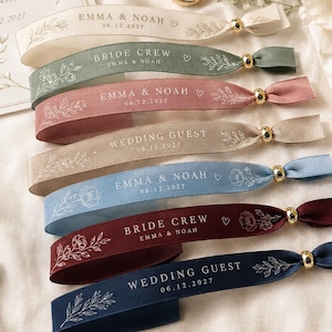 Puede incluir: Una colección de siete pulseras de tela en crema, verde salvia, rosa empolvado, beige, azul claro, burdeos y azul marino. Cada pulsera cuenta con una cuenta dorada, dibujos florales de línea y texto como Emma & Noah, Bride Crew o Wedding Guest.