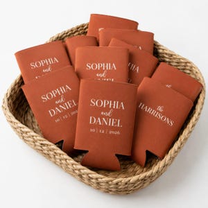 May include: A collection of burnt orange foam can coolers featuring white printed text. Some coolers display the names Sophia and Daniel with the date 10 | 12 | 2026, while others feature the text the Harrisons. The items are arranged in a woven basket.