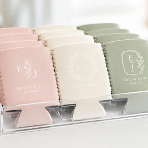 May include: A collection of pink, cream, and sage green foam can coolers arranged in a clear acrylic tray. Each cooler features white printed text with the initials E J, the names Emily & Jacob, and the date 06.14.2025 in various floral designs.