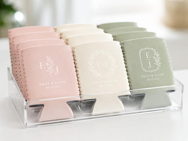 May include: A collection of pink, cream, and sage green foam can coolers arranged in a clear acrylic tray. Each cooler features white printed text with the initials E J, the names Emily & Jacob, and the date 06.14.2025 in various floral designs.