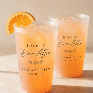 May include: Two frosted plastic cups filled with orange liquid and ice cubes. Each cup features black text reading "HAPPILY Ever After" above a small leaf graphic, followed by "LILY and ETHAN" and the date "05.30.27". An orange slice garnishes the front cup.