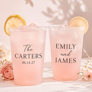 Op de afbeelding: Twee doorzichtige plastic bekers gevuld met roze vloeistof, ijsblokjes en een schijfje citroen. De linkerbeker bevat de tekst "The CARTERS 06.14.27" en de rechterbeker "EMILY and JAMES" in een zwart schreeflettertype.