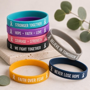 May include: A collection of colorful silicone wristbands with inspirational messages and awareness ribbons. The bands are in various colors, including teal, purple, pink, black, gray, yellow, and navy blue. Messages include "Stronger Together" and "Faith Over Fear."