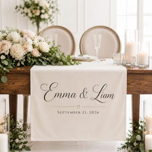 May include: A cream-colored fabric table runner featuring the black printed names Emma & Liam, a small heart icon, and the date SEPTEMBER 21, 2026. The runner is draped over a wooden table decorated with white floral arrangements and candles.