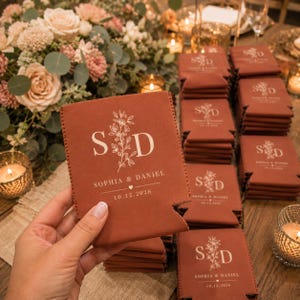 May include: A rust-colored foam can cooler featuring a white printed design with the initials S and D, a floral illustration, the names Sophia & Daniel, a small heart icon, and the date 10.12.2026, with a stack of identical coolers nearby.