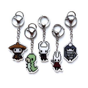 May include: Five pixel art keyrings with silver chains and clasps. The keyrings showcase various characters in different colours, including a brown-hatted figure, a green creature, a white character, a white character in a red cape, and a grey figure.