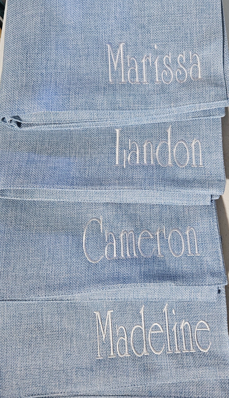 Op de afbeelding: Vier blauwe linnen servetten met witte geborduurde namen: Marissa, Landon, Cameron en Madeline.