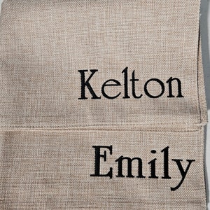 Op de afbeelding: Twee placemats van jute met de namen "Kelton" en "Emily" geborduurd in zwart garen.