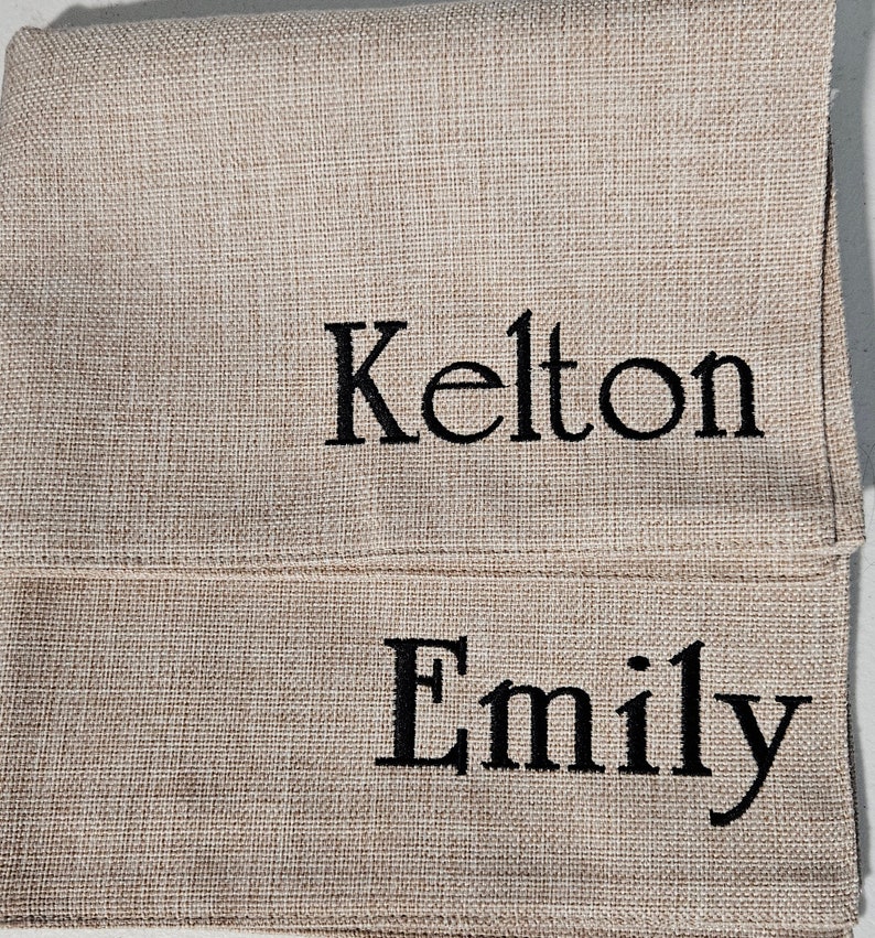 Op de afbeelding: Twee placemats van jute met de namen "Kelton" en "Emily" geborduurd in zwart garen.